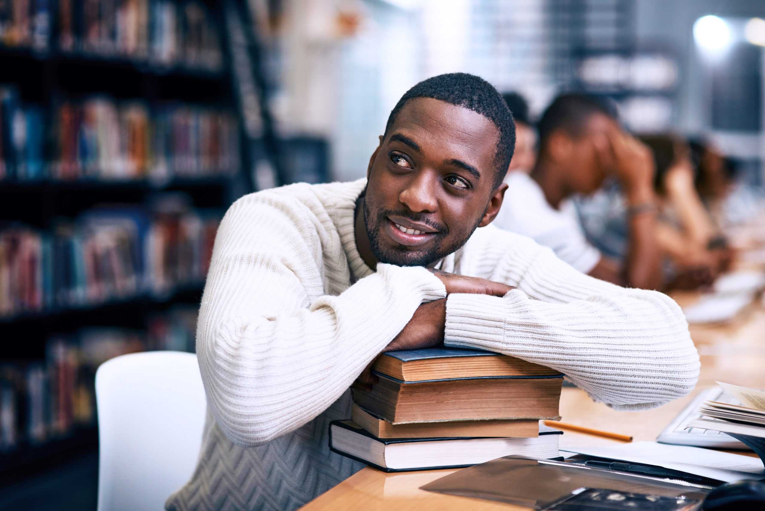 A man dark skin and short, curly hair rests his hands on a pile of books in a library.